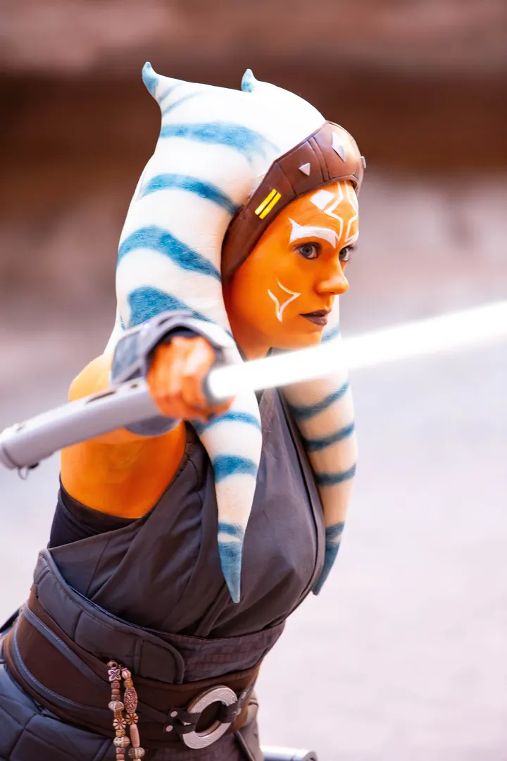 Actress as live-action Ahsoka Tano with a lightsaber on the set of Star Wars Rendezvous, wearing a Wretched Hive headpiece with blue-striped lekku